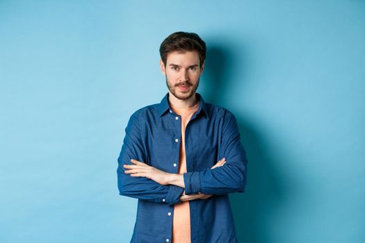 Confident young man feeling like professional, cross arms on chest and smiling sassy, standing on blue background.