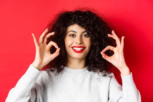 Close-up portrait of smiling woman with curly hair and red lips, showing okay gesture and looking satisfied, praise good product, standing on red background.