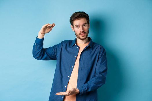 Modern guy showing big size, shaping large object and looking impressed at camera, standing on blue background.