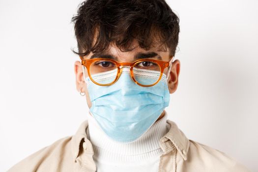 Headshot of young guy in glasses and sterile medical mask looking cheerful and motivated, standing on white background. Social distancing and coronavirus concept.