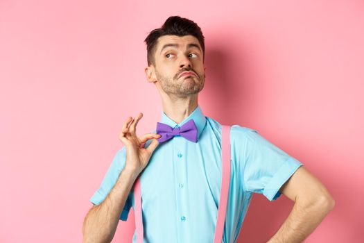 Image of funny man raising chin up and adjusting bow-tie, standing confident and sassy on pink background, looking left.