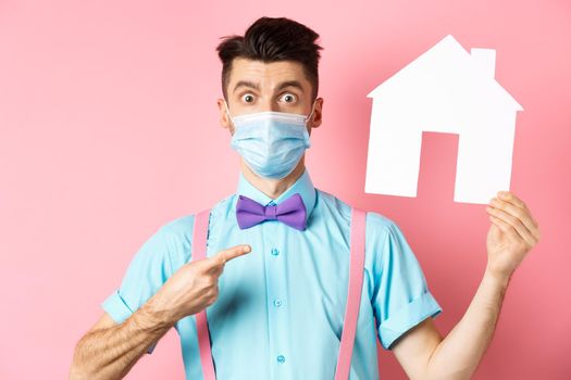 Covid, pandemic and real estate concept. Amazed man in face mask pointing at paper house cutout, standing impressed on pink background.