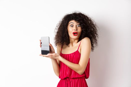 Excited woman with curly hair, makeup and red dress, gasping amazed, showing empty smartphone screen with amazed face, standing against white background.