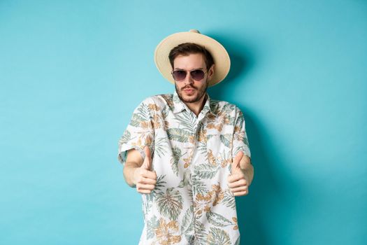 Handsome tourist enjoying summer holiday, wearing sunglasses and hawaiian shirt, showing thumbs-up in approval, standing on blue background.