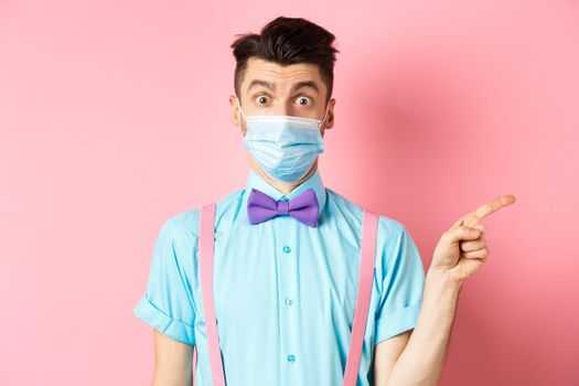 Coronavirus, healthcare and quarantine concept. Man looking surprised in medical mask, asking question and pointing right, curious about promo offer, standing on pink background.