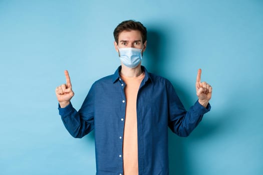 Covid-19 and lifestyle concept. Handsome young man in face mask looking healthy and happy, pointing fingers up at empty space, blue background.