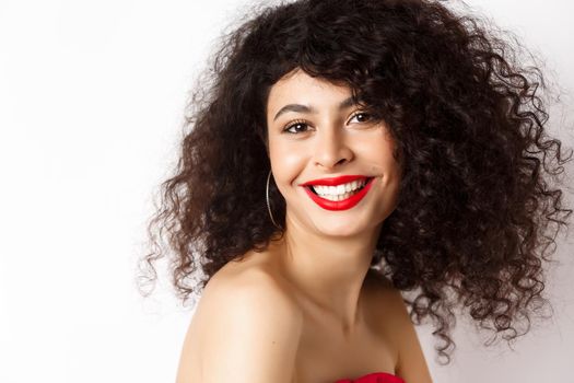Close-up of cheerful smiling woman with red lipstick and curly hairstyle, looking happy, standing over white background.