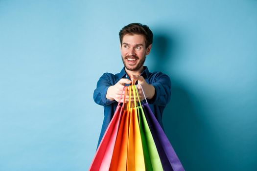 Handsome happy guy smiling, stretch out hands with shopping bags and looking aside at empty space, standing on blue background.