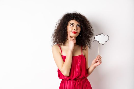 Thoughtful girl in red dress, holding small cloud and looking up with pleased smile, imaging things, standing over white background.