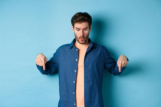 Impressed caucasian male model in casual clothes, say wow, looking and pointing fingers down at logo, standing on bllue background.