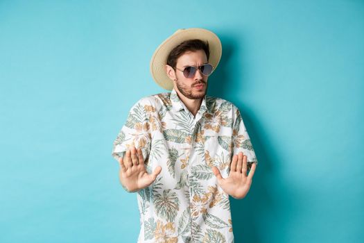 Alarmed tourist asking to stay away, step back from something cringe, showing rejection gesture, standing in straw hat and hawaiian shirt, blue background.