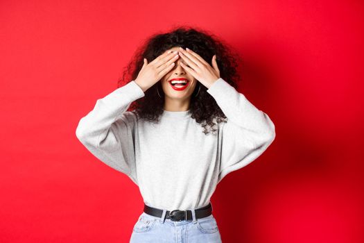 Happy beautiful woman with curly hair and red lips, covering eyes with hands and waiting surprise, smiling excited, standing against red background.