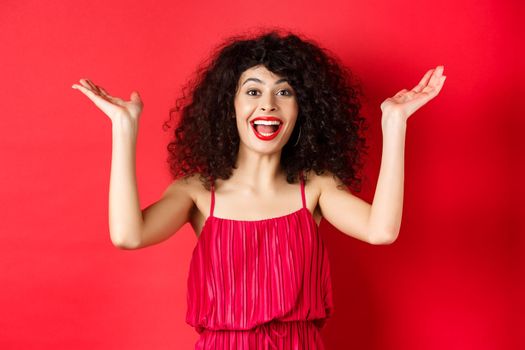 Excited beautiful woman raising hands up and scream with happiness and joy, applause with rejoice, standing on red background.
