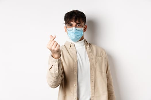 Covid-19, health and real people concept. Cute young man in medical mask showing finger heart and look at camera, standing on white background.