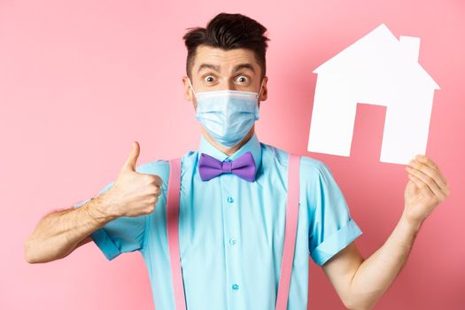 Covid, pandemic and real estate concept. Satisfied agency client showing thumb up and paper house cutout, wearing medical mask, standing over pink background.