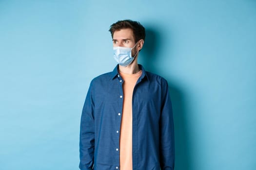 Covid-19 and healthcare concept. Shocked young man in medical mask looking aside at empty space on blue background.