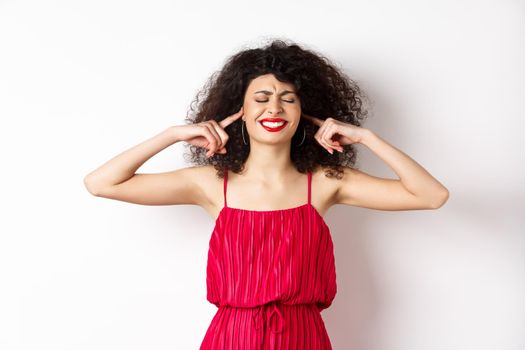 Annoyed woman with curly hair and red dress, shut eyes and ears, block annoying sound, clench teeth bothered, complain on loud music or neighbours, standing on white background.