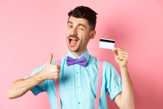 Shopping concept. Festive guy in bow-tie winking at camera, showing thumbs up with plastic credit card, recommending good shop offer, promote bank, standing on pink background.