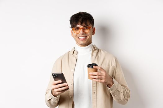 Natural handsome man in glasses drinking coffee from paper cup and using cellphone, smiling satisfied at camera, white background.