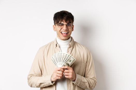 Handsome young man winking and showing dollar bills, holding fan of money banknotes and smiling pleased, making money, standing on white background.