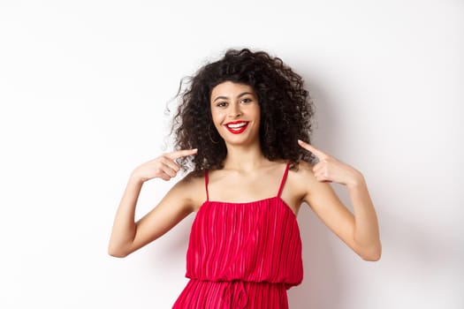 Beautiful woman with curly hairstyle, makeup and red dress, pointing at herself, smiling with white teeth, standing on white background.
