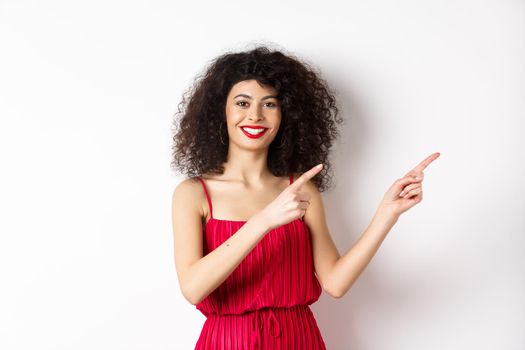 Cheerful female model in fashionable red dress, smiling and pointing fingers right at logo, standing over white background.