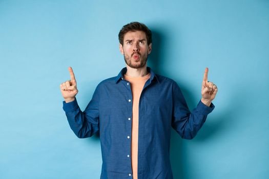 Confused young man frowning, pointing fingers up and looking displeased, standing on blue background. Copy space