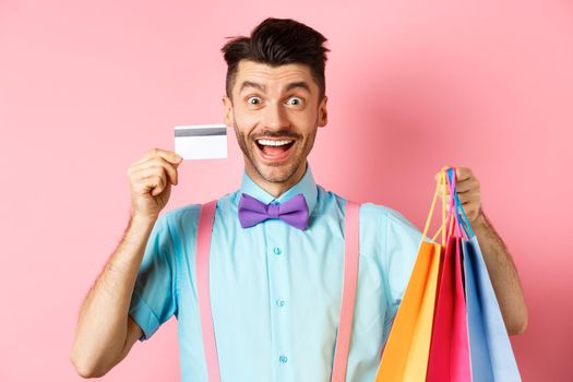 Excited man showing plastic credit card and shopping bags, smiling amazed while buying gifts, standing on pink background.
