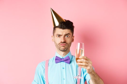 Holidays and celebration concept. Troubled young man in party hat, frowning with doubtful face, raising glass of champagne perplexed, standing on pink background.