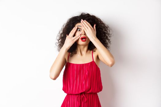 Excited curly-haired girl in dress, cover eyes with hands and peek through fingers, gasping shocked at camera, standing on white background.