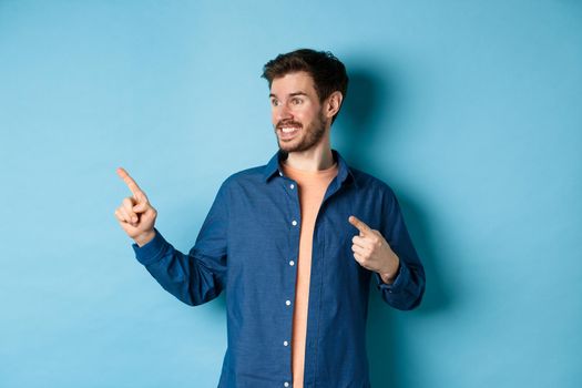 Smiling handsome guy pointing and looking left at empty space, showing logo, standing on blue background.