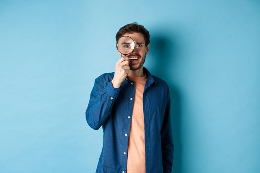 Cheerful young man found something good, smiling and looking through magnifying glass, standing on blue background.