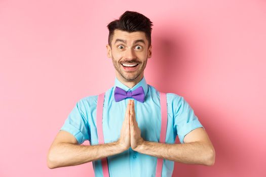 Image of smiling young man asking please, looking grateful at camera, Guy in bow-tie say thank you, standing over pink background.