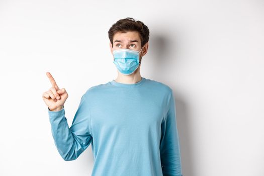 Coronavirus, health and quarantine concept. Curious guy in medical mask, pointing and looking at upper left corner banner, standing on white studio background.