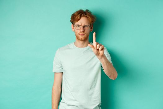 Angry and serious young man with red hair, wearing glasses, showing stop gesture, telling no, shaking finger with disapproval, standing over mint background.