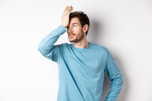 Annoyed young man with beard, roll eyes and making facepalm, pissed-off with something, standing irritated on white background.