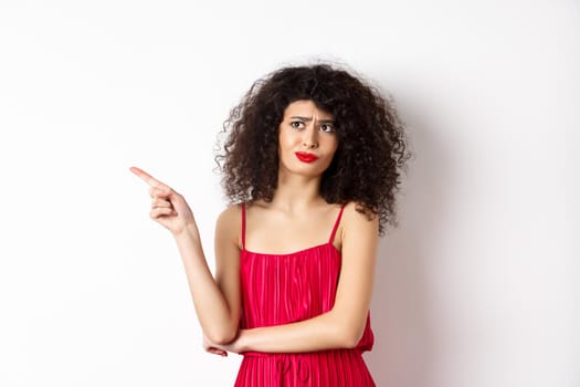 Disappointed and skeptical young woman with curly hair, wearing red dress, grimacing and pointing finger left at logo, standing over white background.
