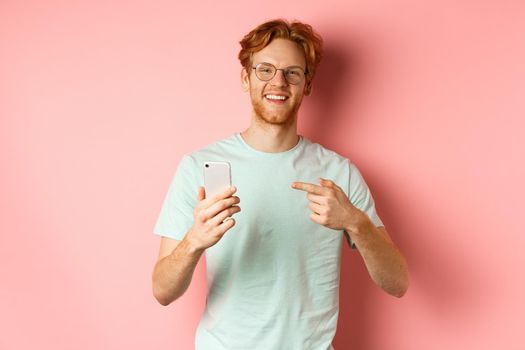 Young man with red hair and beard, wearing t-shirt and glasses, smiling while pointing finger at smartphone, recommend online promotion, pink background.