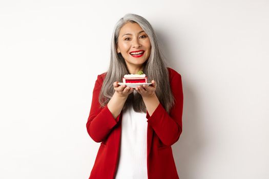 Beautiful asian grandmother holding piece of cake and smiling, standing in red blazer over white background.