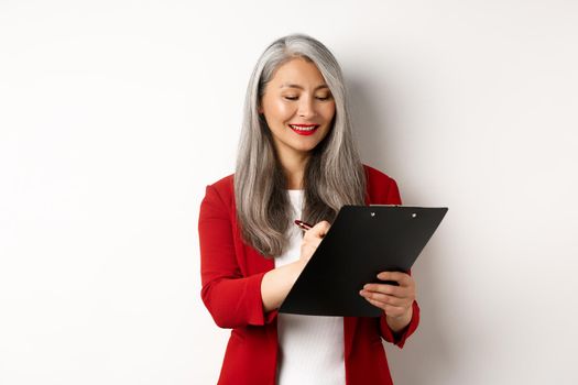 Successful asian senior businesswoman with grey hair taking notes on clipboard, inspecting enterprise, standing over white background.