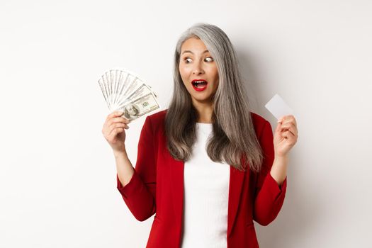 Surprised asian middle-aged woman with grey hair, looking at money and holding credit card, earning cash, standing over white background.