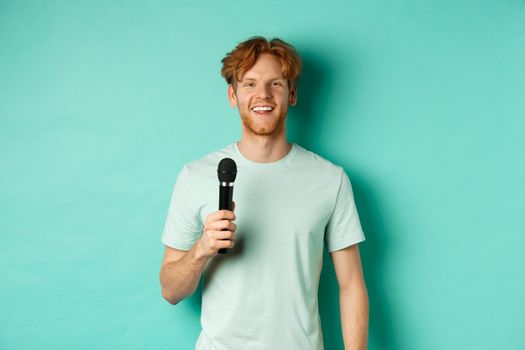 Young redhead man with beard, wearing t-shirt, holding microphone and making speech, singing karaoke, standing over mint background.