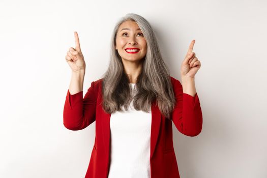 Happy asian lady in red blazer and makeup, looking and pointing fingers up, showing special offer, standing over white background.