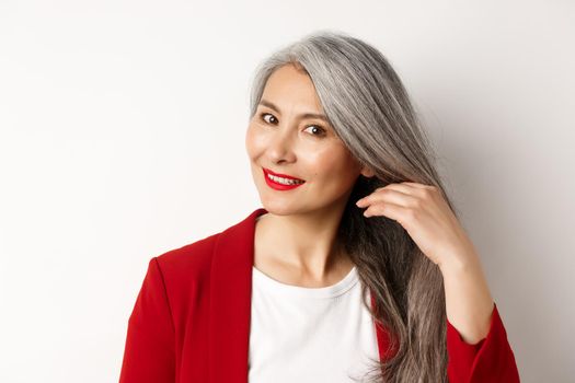 Beauty and haircare concept. Close up of elegant asian senior woman showing shiny and healthy grey hair, smiling and looking aside, white background.