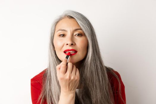 Beauty and fashion concept. Stylish asian mature woman with grey hair, looking in mirror and apply red lipstick, standing over white background.