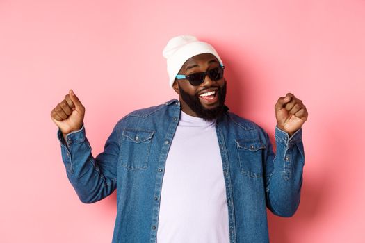 Happy and carefree Black man celebrating, dancing and having fun, wearing beanie and sunglasses, standing over pink background.