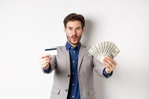 Happy successful businessman in suit showing money and credit card, smiling at camera, white background.