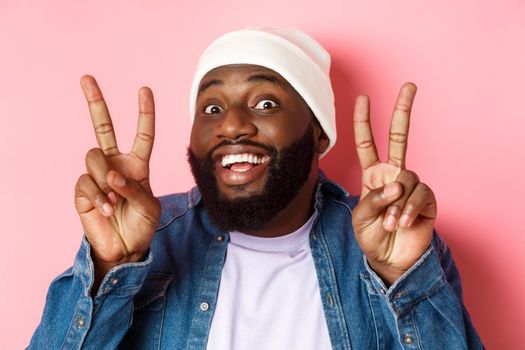 Close-up of cheerful african-american hipster in beanie smiling, showing peace signs, standing happy against pink background.