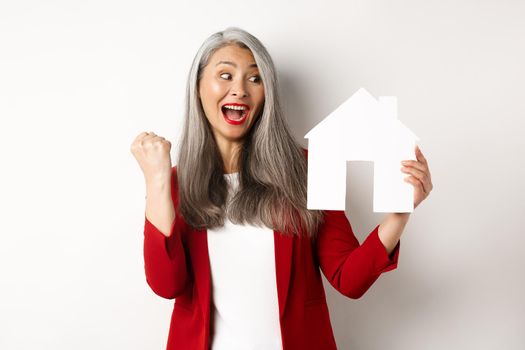 Cheerful asian senior woman buying house, scream of joy and making fist pump while showing paper house cutout, standing over white background.