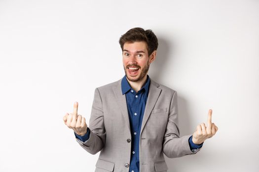 Rude ignorant guy in business suit showing middle fingers and tongue, smiling while mocking people, fuck you gesture, standing on white background.